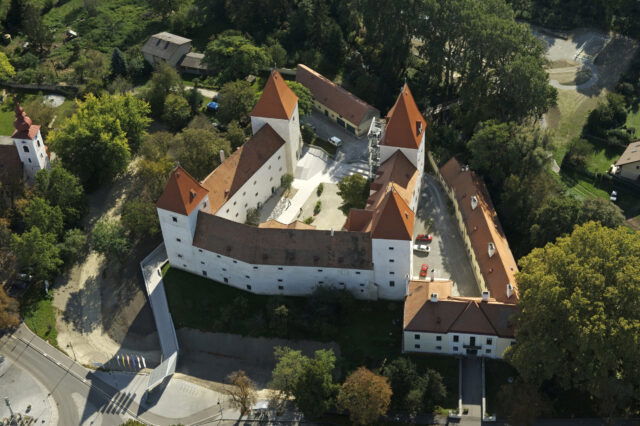 Luftaufnahme von Schloss Orth: Weiße Schlossanlage mit vier markanten roten Spitztürmen, umgeben von Bäumen.