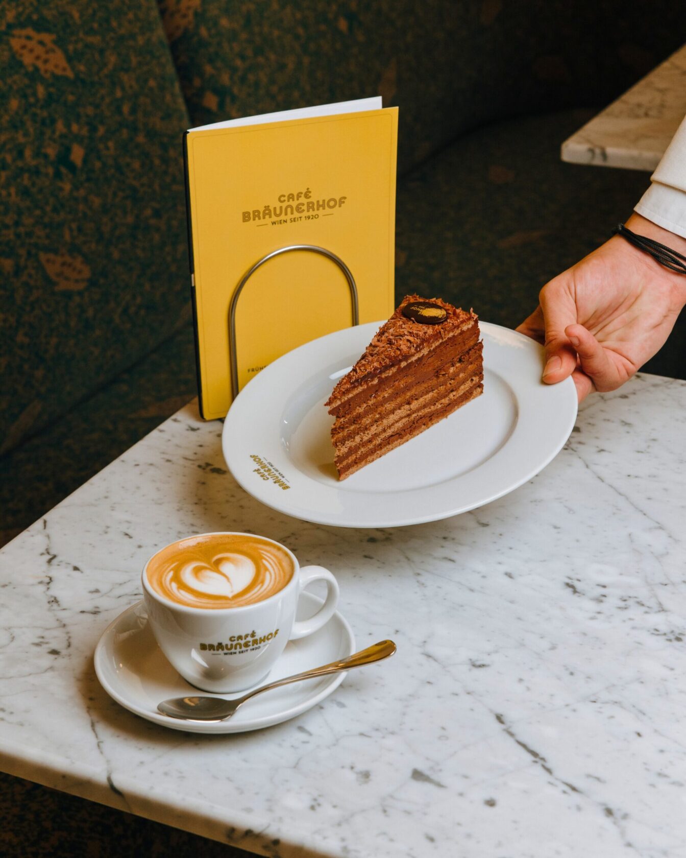 ein Kellner stellt ein Stück Kuchen auf einen Tisch ein historischer Zeitungsständer steht im Café Bräunerhof in Wien