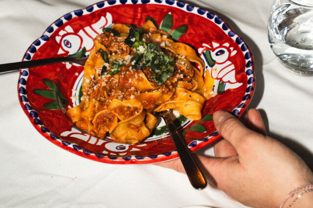Pasta auf einem bunten Teller, der in der Hand gehalten wird. Seitlich davon steht Parmesan am Tisch.