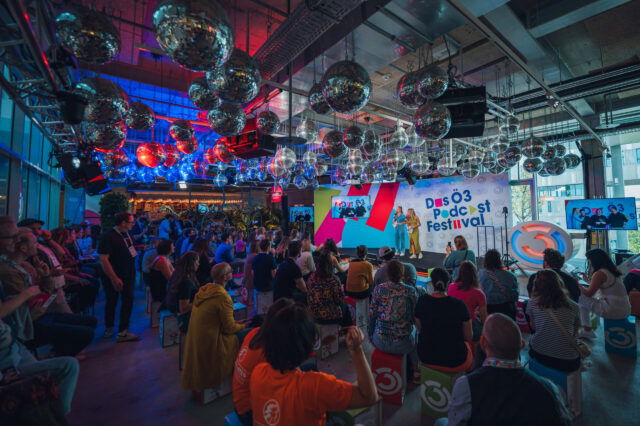 Das Ö3 Podcast Festival Bühne: Zwei Moderatorinnen vor einer weißen Wand mit buntem Veranstaltungslogo; rechts ein großes, beleuchtetes Ö3-Logo. Zuschauerraum: Gut gefüllt, Publikum sitzt auf bunten Hockern. Moderne Event-Location mit industrieller Decke, dicht behängt mit zahlreichen spiegelnden Discokugeln.Stimmungsvolles blaues und rotes Licht.