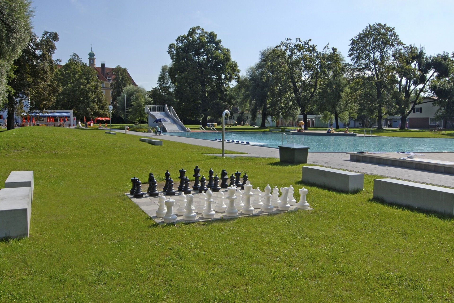 Ein Schachbrett mit großen Spielfiguren auf einer grünen Wiese mit Freibad in München im Hintergrund.