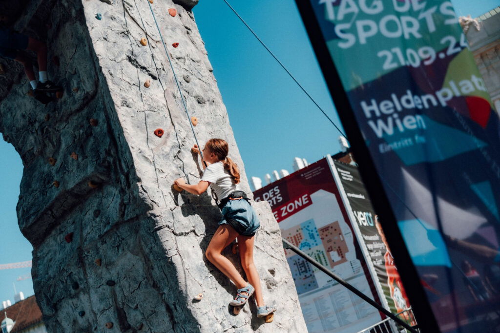 Ein junges Mädchen probiert die Mitmachstation „Klettern“ beim Tag des Sports am Heldenplatz.