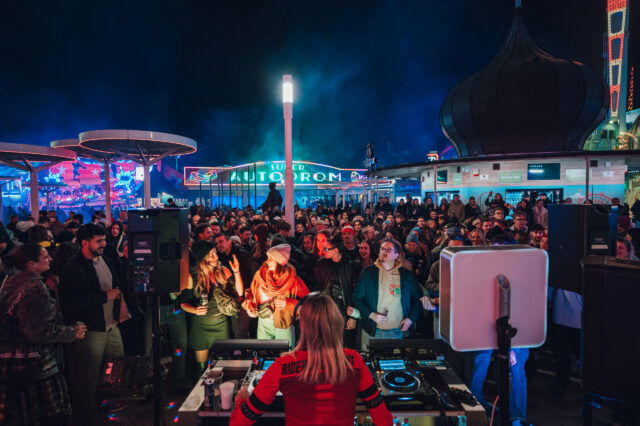 Menschen stehen bei Nacht im Wiener Prater und tanzen, im Vordergrund ein DJ Pult