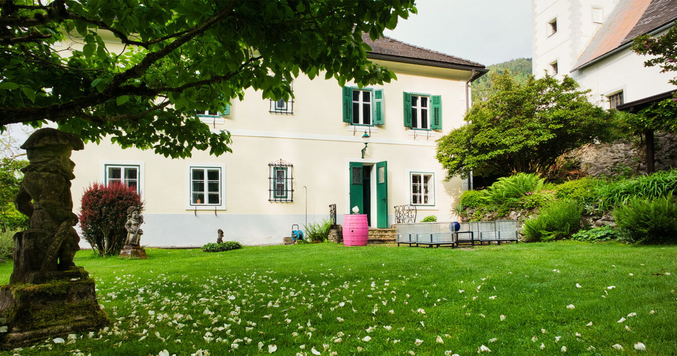 Idyllisches gelbes Landhaus mit grünen Fensterläden, gepflegtem Garten, Steinstatue und rosa Weinfass.