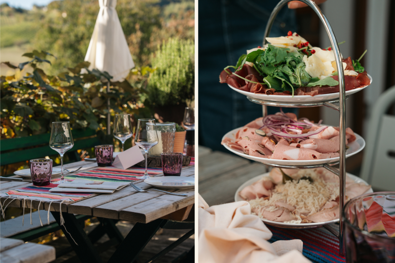 Gedeckter Holztisch auf der Terrasse eines steirischen Buschenschanks mit Weingläsern und einer dreistöckigen Etagere voll regionaler Jausen-Spezialitäten.