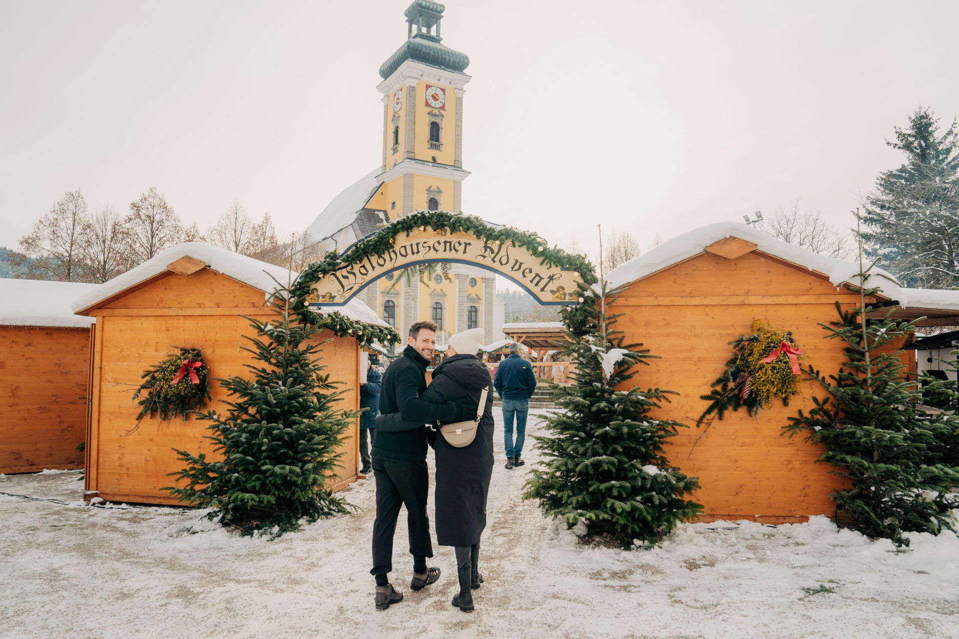 Ein Paar steht am Eingang zum Adventmarkt in Waldhausen, direkt vor der imposanten Stiftkirche. Die festlich geschmückte Kulisse und die Vorfreude auf das Erkunden des Marktes spiegeln sich in ihren strahlenden Augen wider.