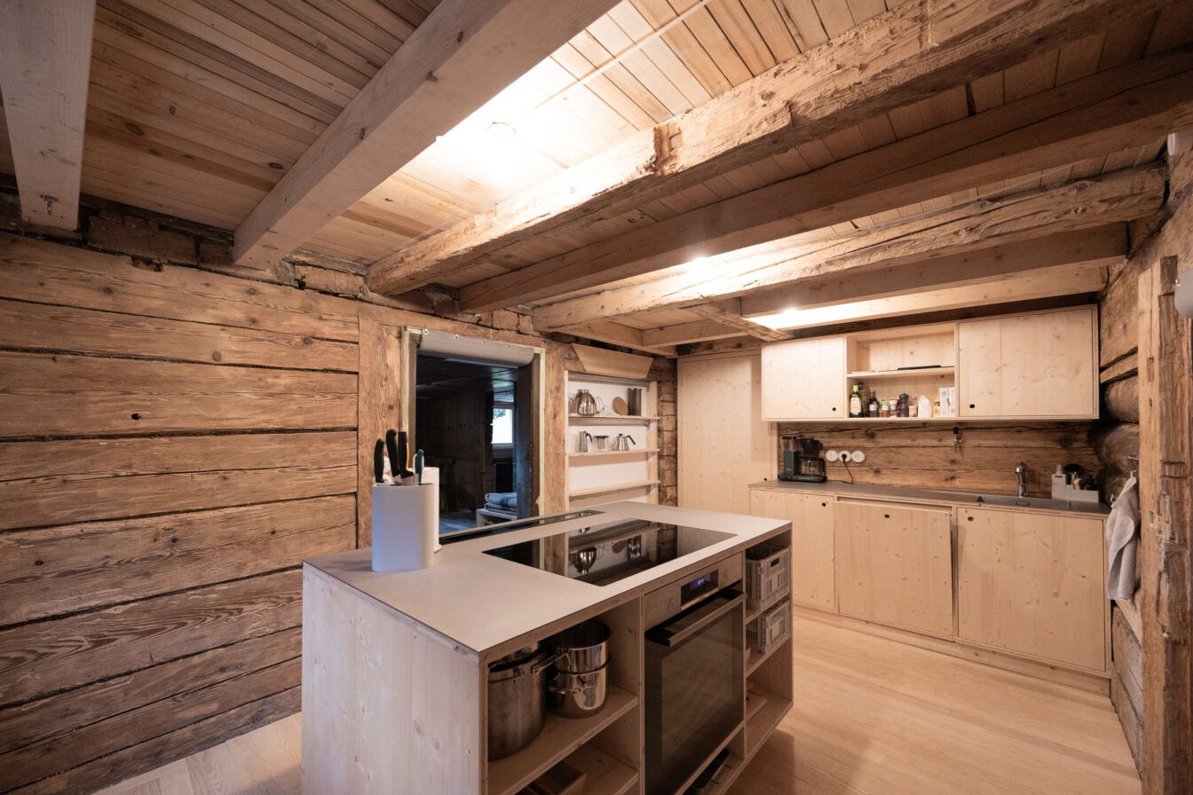Rustikale Küche in einem Holzhaus mit freiliegenden Holzbalken, Kücheninsel, modernem Kochfeld und hellen Holzfronten.