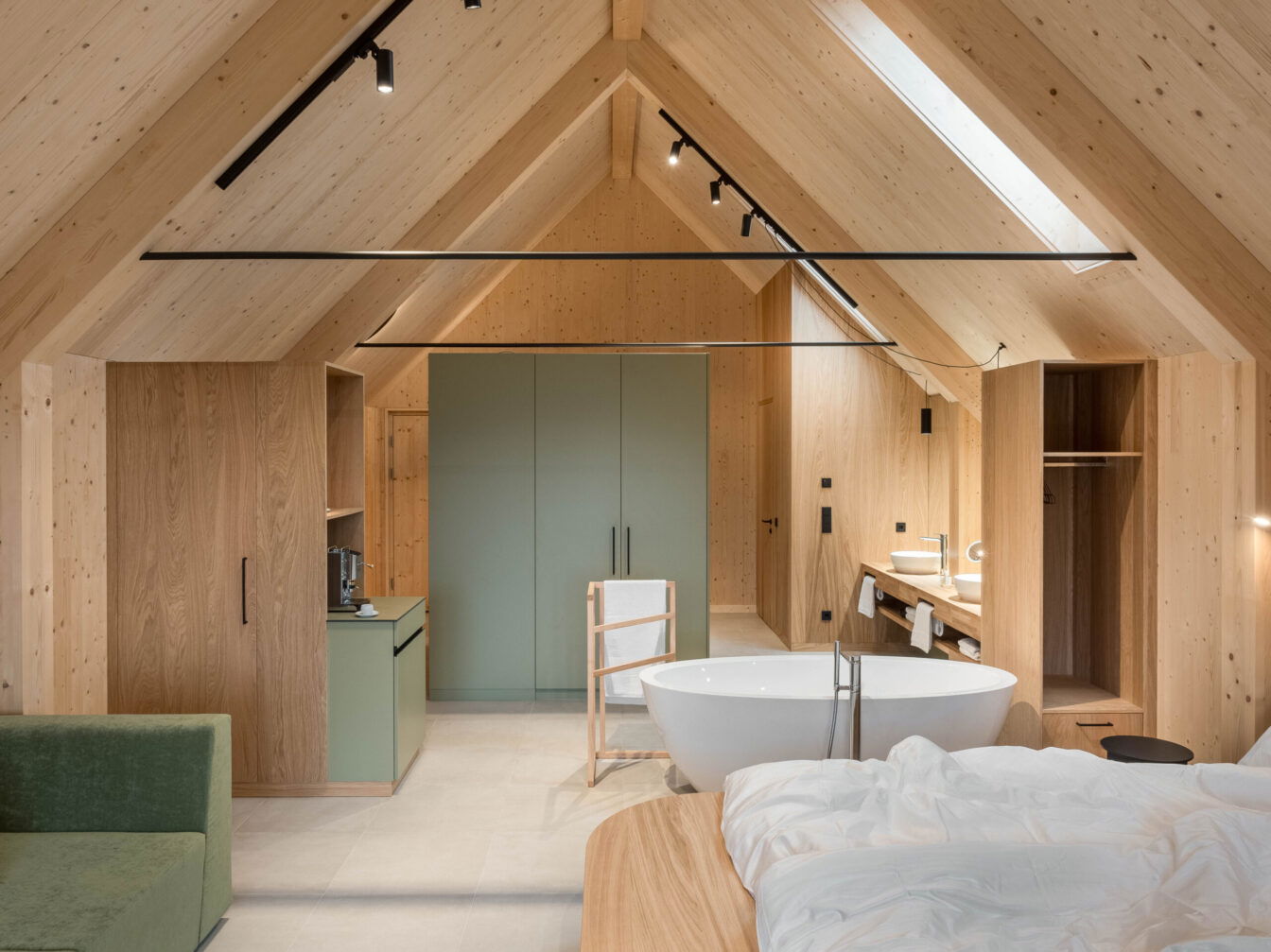 Moderner Open-Space-Schlafraum mit Sichtdachstuhl aus hellem Holz, freistehender Badewanne, salbeigrünen Schränken und offenem Badbereich.