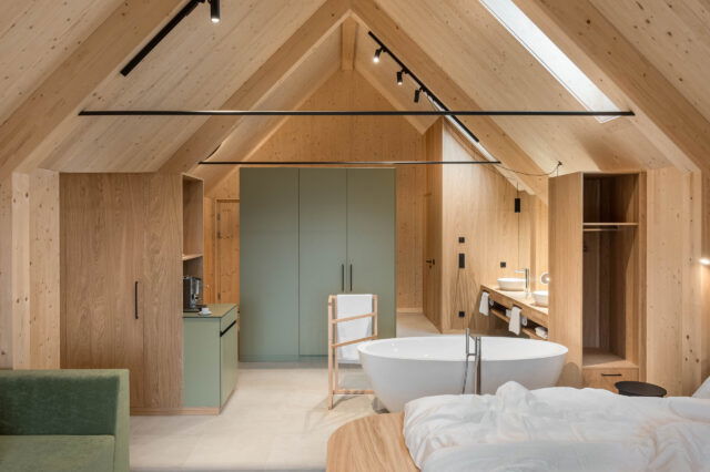 Moderner Open-Space-Schlafraum mit Sichtdachstuhl aus hellem Holz, freistehender Badewanne, salbeigrünen Schränken und offenem Badbereich.