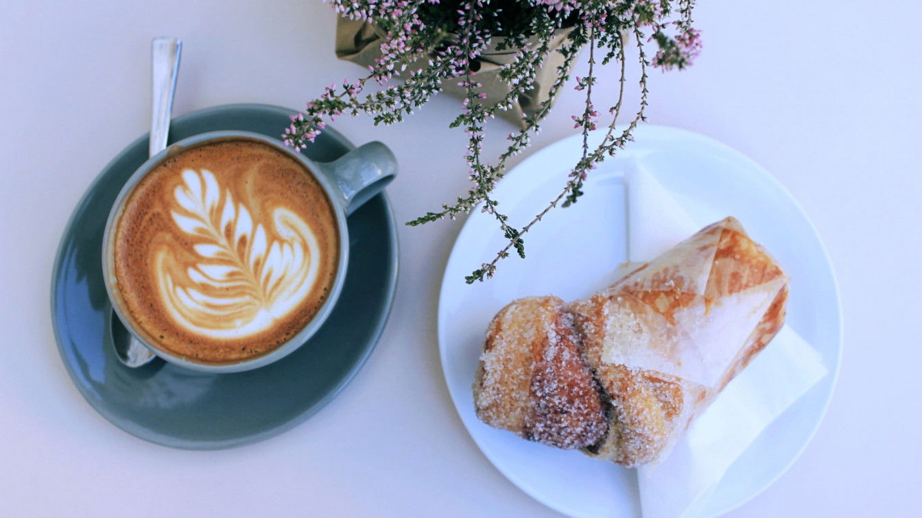 Vogelsicht auf einem creme-weißen Tisch. Links im Bild ein Kaffee mit Latte Art in grauem Geschirr, rechts auf einem weißen Teller und auf einer Serviette ein Cruffin mit Zimt und Zucker.